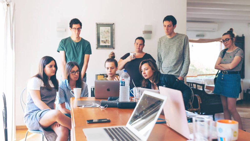 réunion de travail avec 6 personnes autour d'un bureau en bois de couleur miel. Il y a 3 hommes debout et 3 femmes assises. Les 6 personnes regardent le même ordinateur de marque apple. On peut voir sur le bureau 2 autres ordinateurs portables ouverts mais éteints, un téléphone portable, une bouteille d'eau, un mug, un bloc notes avec une couverture en cuir noir