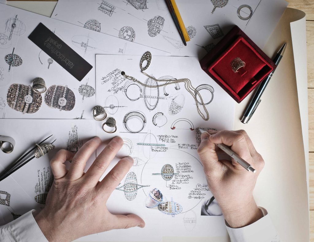 deux mains blanches, celle de gauche est ouverte et tient les feuilles, la main droite tient un stylo et dessine sur une des feuilles. plusieurs feuilles sont éparpillées sur la table, sur lesquelles on voit des bijoux dessinés : essentiellement des bagues. une règle noire se trouve en haut à gauche. un étui rouge foncé se trouve en haut à droite de la photo. un stylo noir est posé à droite de l'étui rouge foncé et un crayon de papier est placé à gauche de l'étui rouge foncé.
